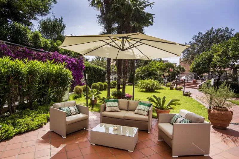 Salon de jardin avec canapés et table basse sous un parasol, entouré de végétation luxuriante.