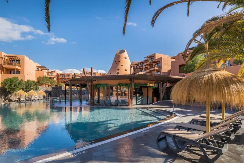 Piscine turquoise bordée de transats devant un bâtiment aux tons ocre et une végétation luxuriante.