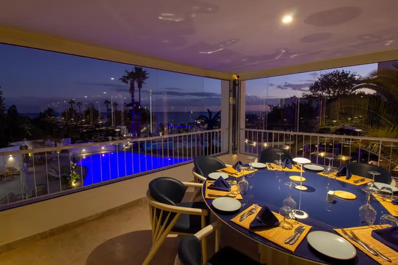Une table ronde est dressée pour un repas avec vue sur une piscine illuminée et la mer au crépuscule.