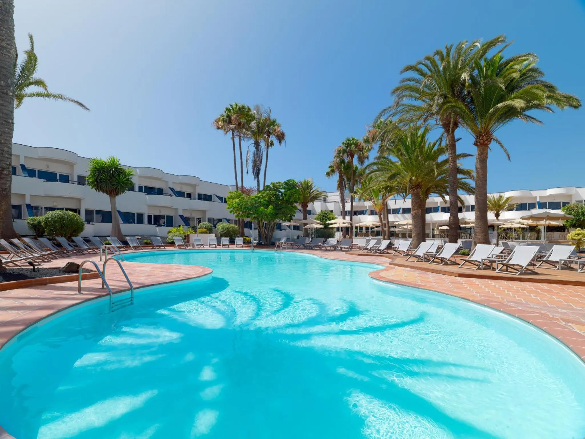 Une piscine turquoise bordée de transats et de palmiers devant un bâtiment blanc.