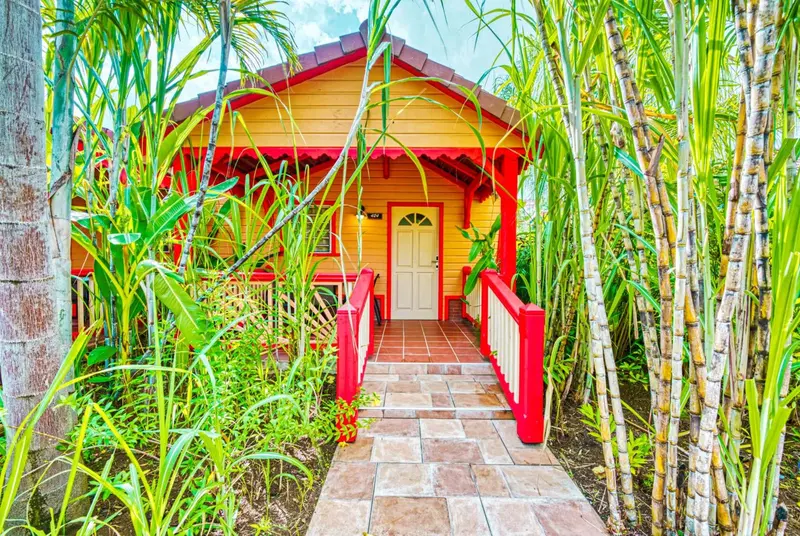 Une petite maison jaune avec un toit rouge et une porte blanche est accessible par un pont en bois et des marches.