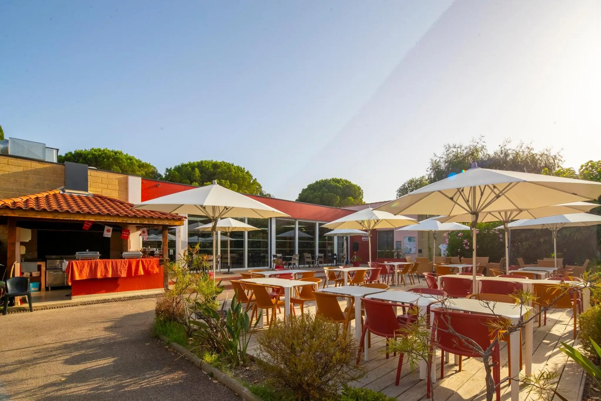 Terrasse ombragée avec tables et chaises, devant un bâtiment bas.