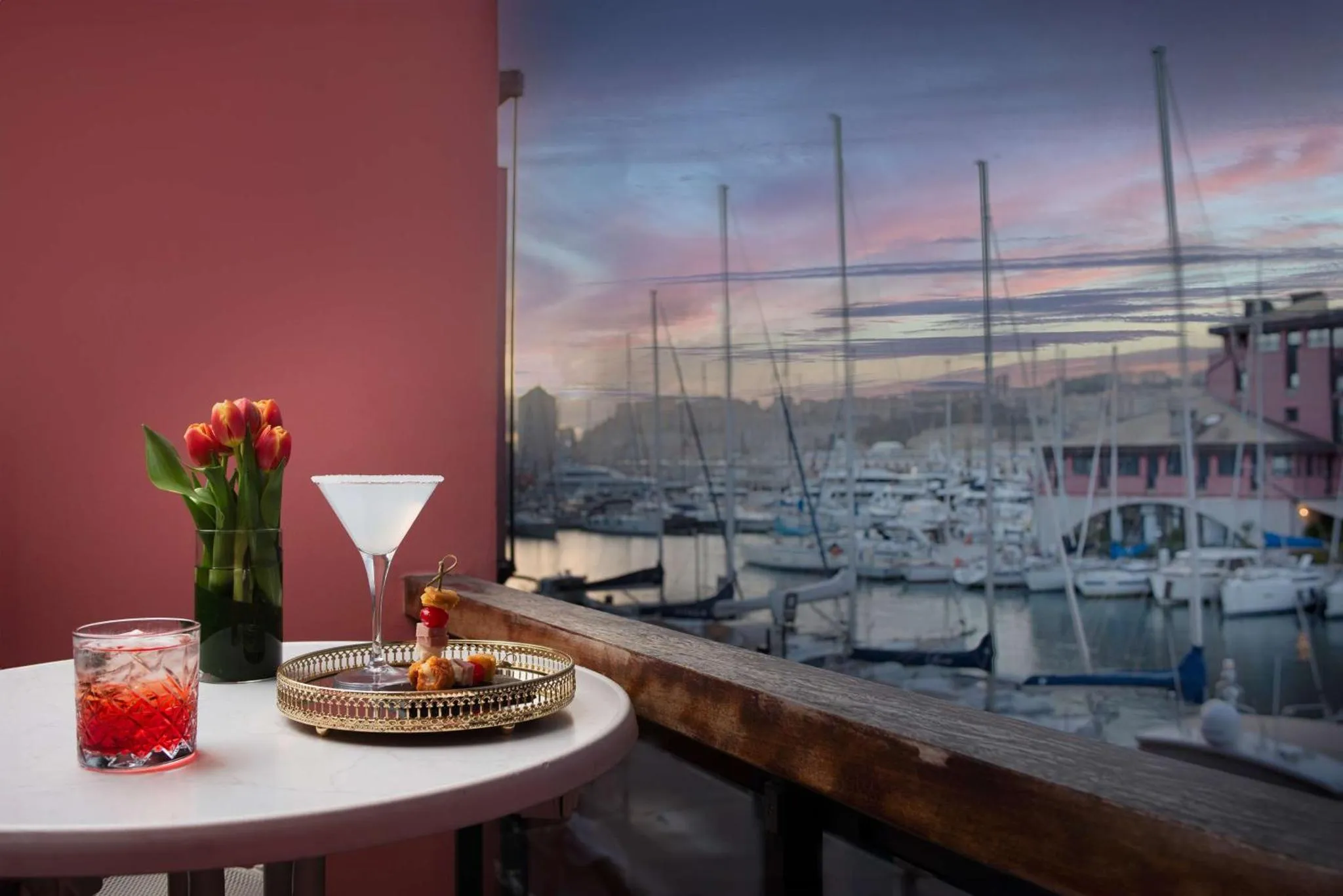 Une table avec des boissons et des amuse-gueules sur une terrasse donnant sur un port de plaisance au coucher du soleil.