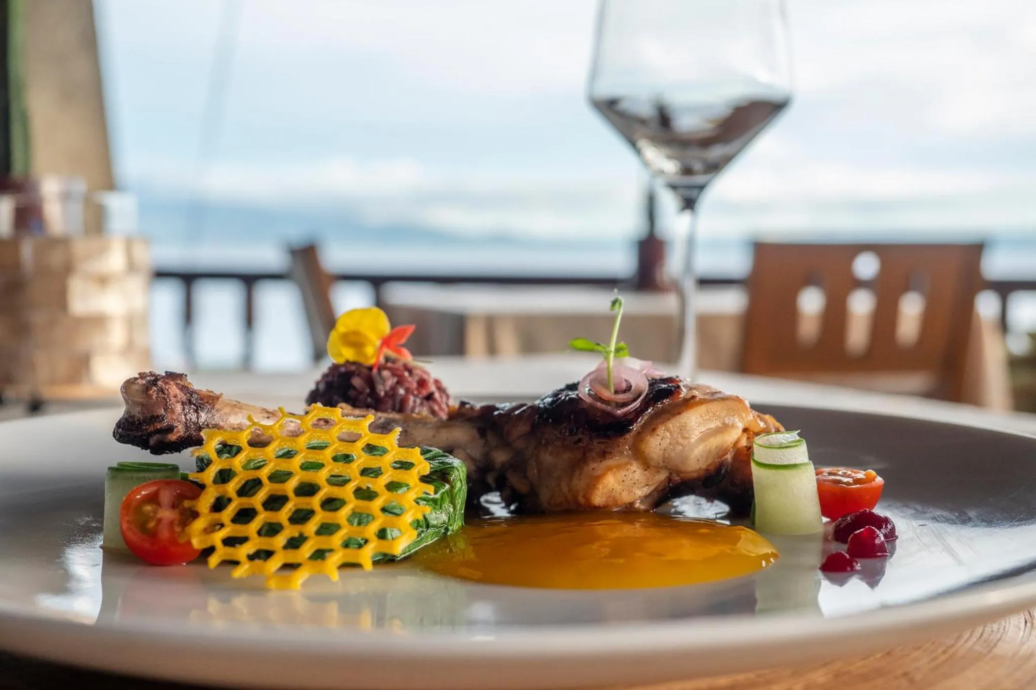 Assiette de viande grillée décorée, accompagnée d'un verre de vin et vue sur la mer.
