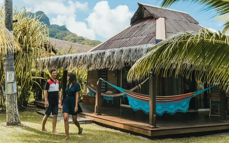 Un couple se promène devant un bungalow sur pilotis avec un hamac.