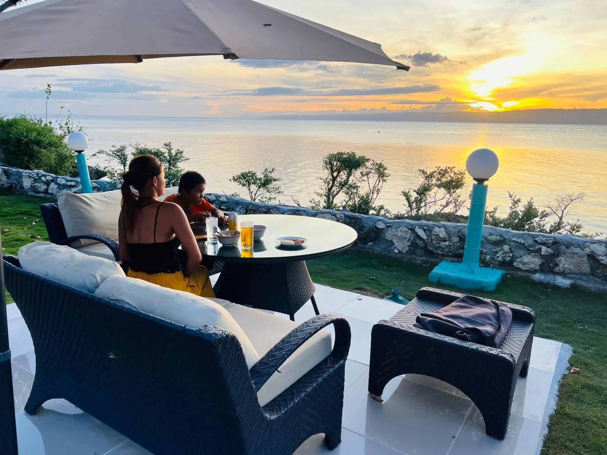 Deux personnes sont assises à une table en terrasse, face à la mer au coucher du soleil.