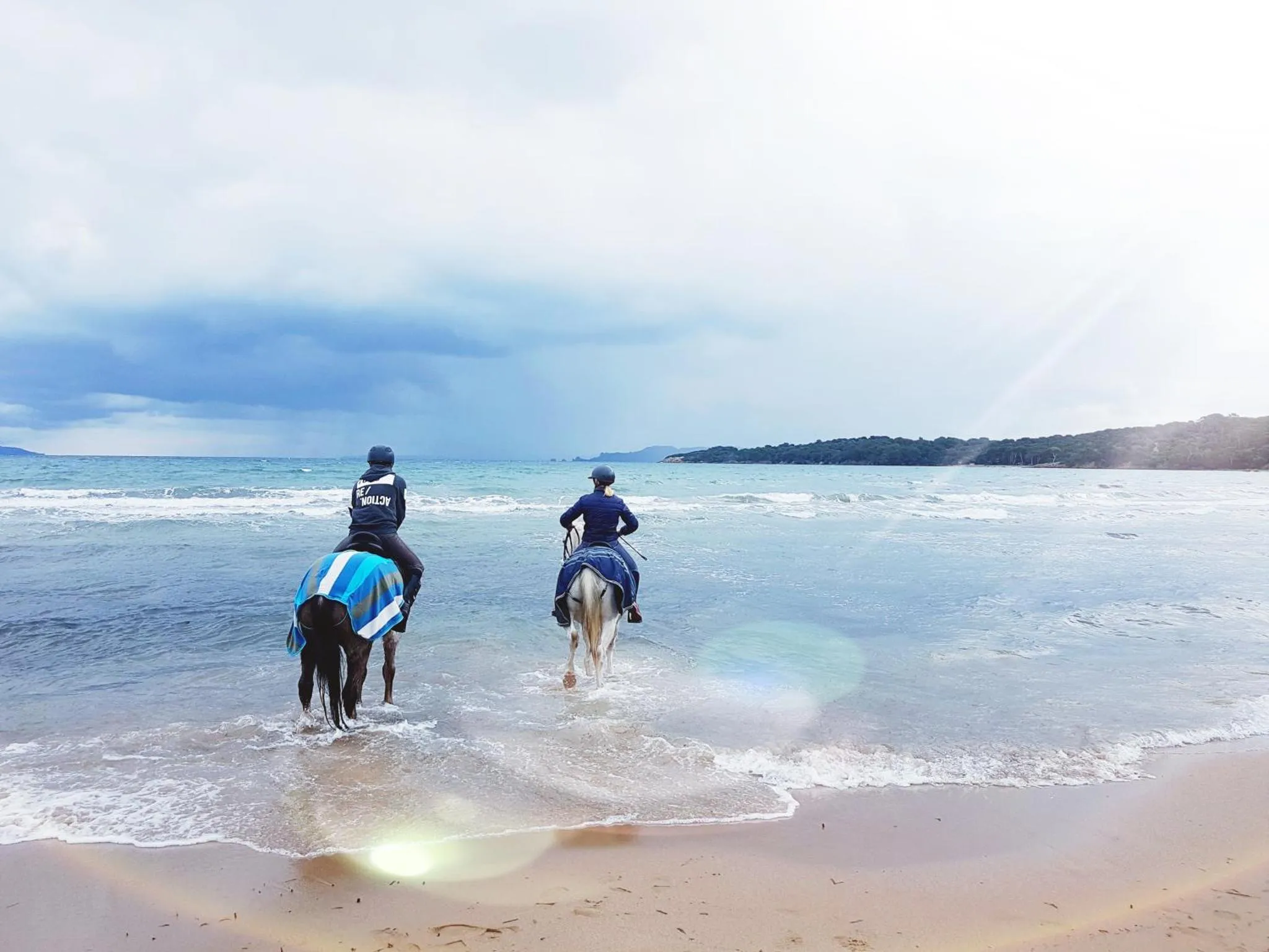 Deux cavaliers s'éloignent sur une plage de sable clair face à la mer.