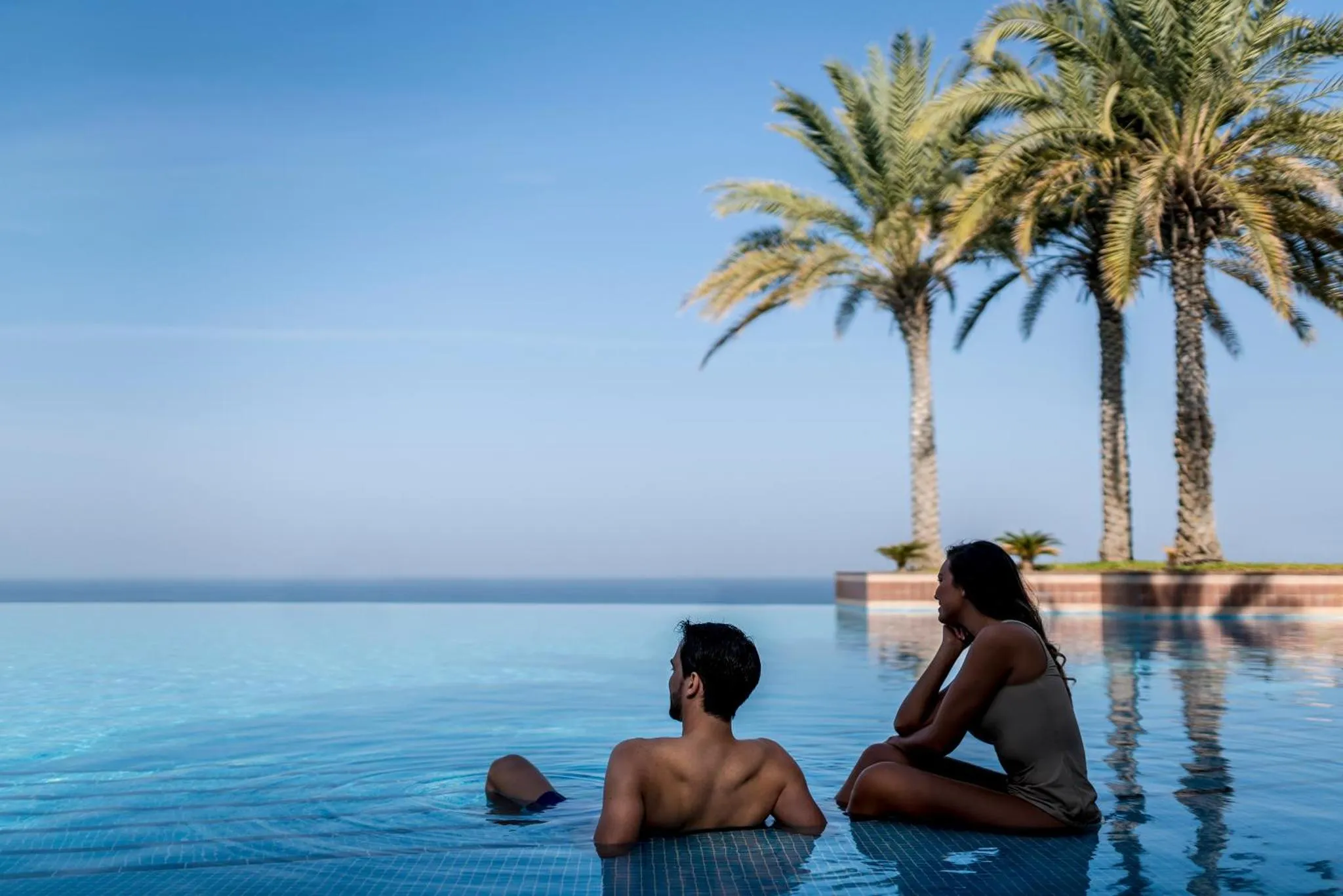 Deux personnes se détendent au bord d'une piscine à débordement face à la mer.