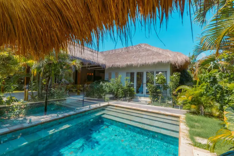 Une piscine turquoise bordée d'une terrasse ensoleillée devant un bâtiment à toit de chaume.