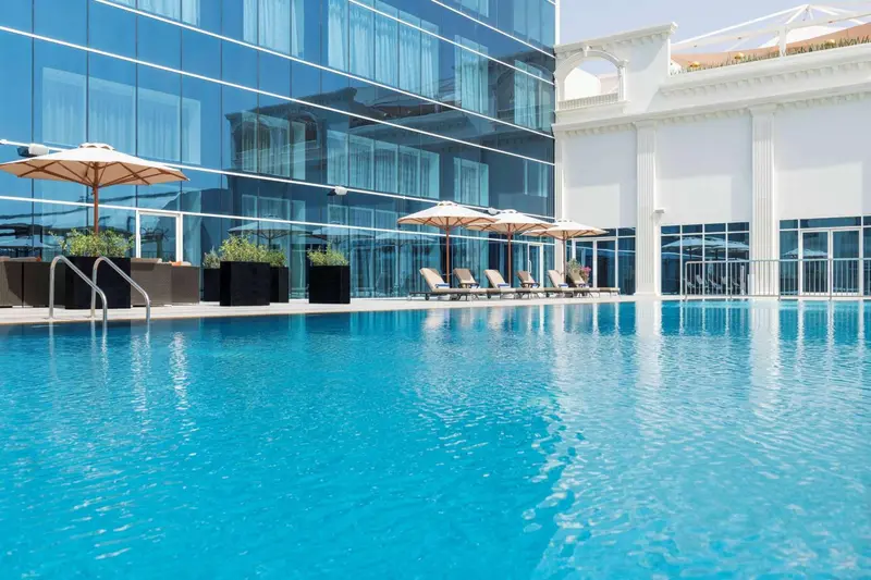 Une piscine bleu clair avec des transats et des parasols blancs, devant un bâtiment moderne à la façade vitrée.