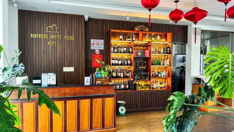 Un comptoir d'accueil en bois clair dans un hall d'hôtel décoré de plantes et de bouteilles.