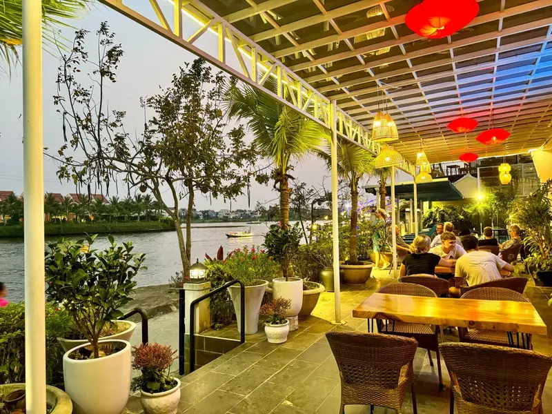 Une terrasse ombragée avec des tables et des chaises en osier, donnant sur une rivière au coucher du soleil.