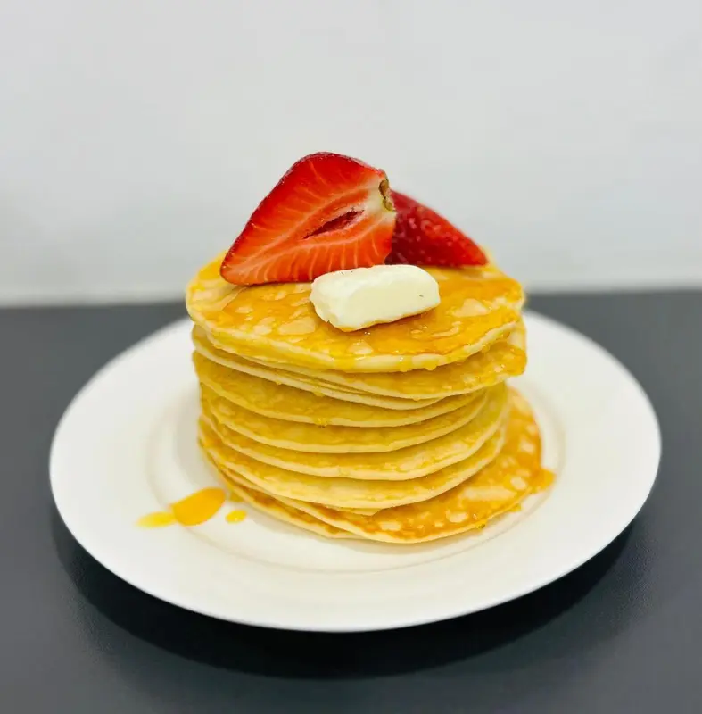 Une pile de crêpes dorées, nappées de sirop et garnies d'une fraise et d'une noix de beurre, est présentée sur une assiette blanche.