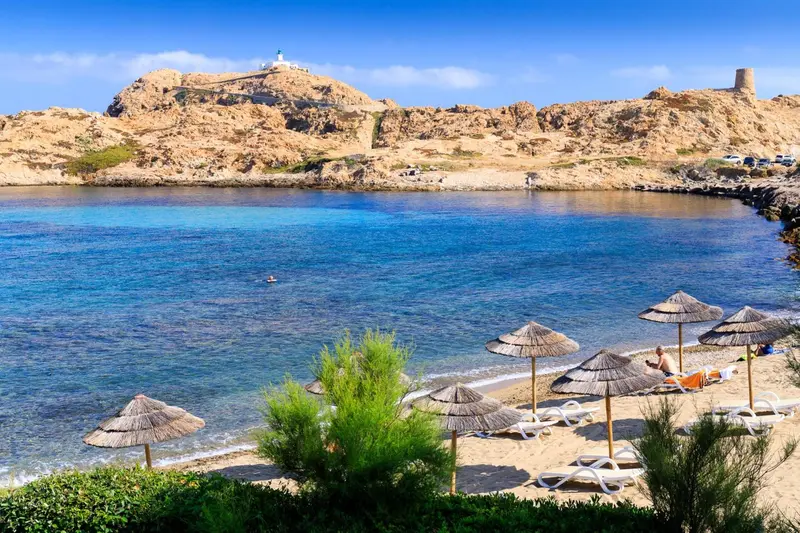 Une plage de sable avec des parasols et des transats, face à une crique turquoise et une colline rocheuse.