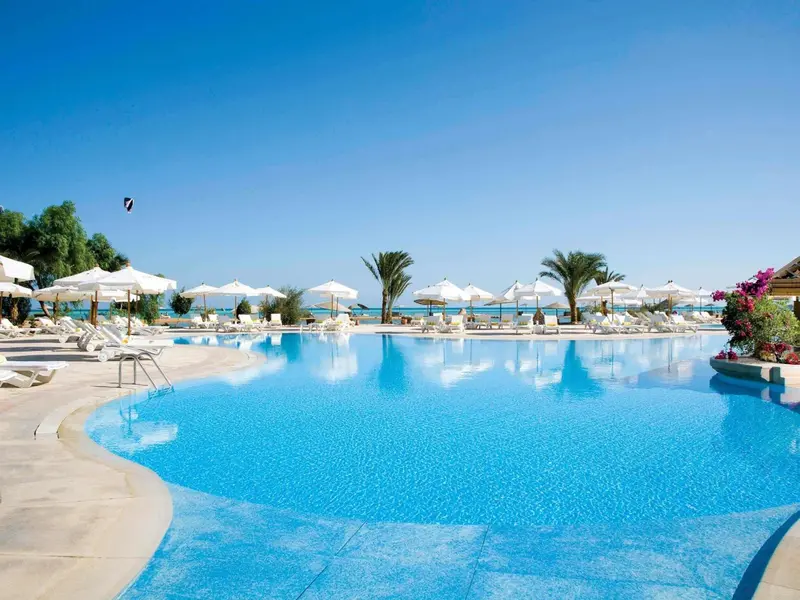 Une piscine turquoise bordée de transats blancs sous des parasols, avec la mer en arrière-plan.