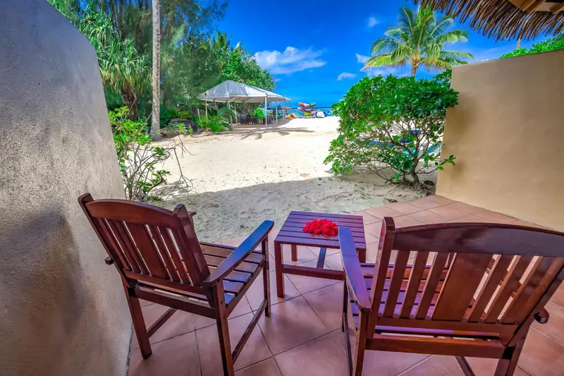 Deux fauteuils en bois et une petite table basse sur une terrasse carrelée donnant sur une plage de sable blanc.