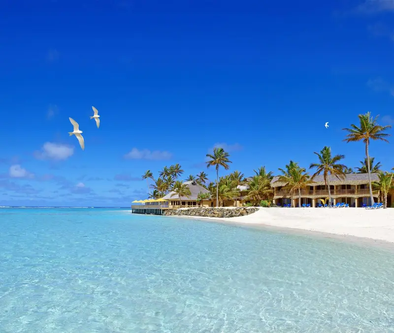 Plage de sable blanc bordée d'eau turquoise et de végétation tropicale sous un ciel bleu.