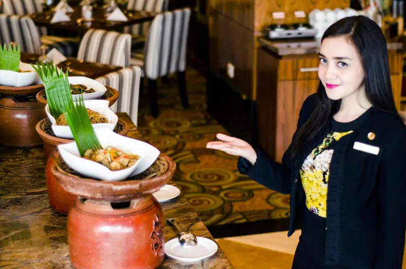 Une employée souriante présente un buffet de plats variés dans une salle lumineuse.