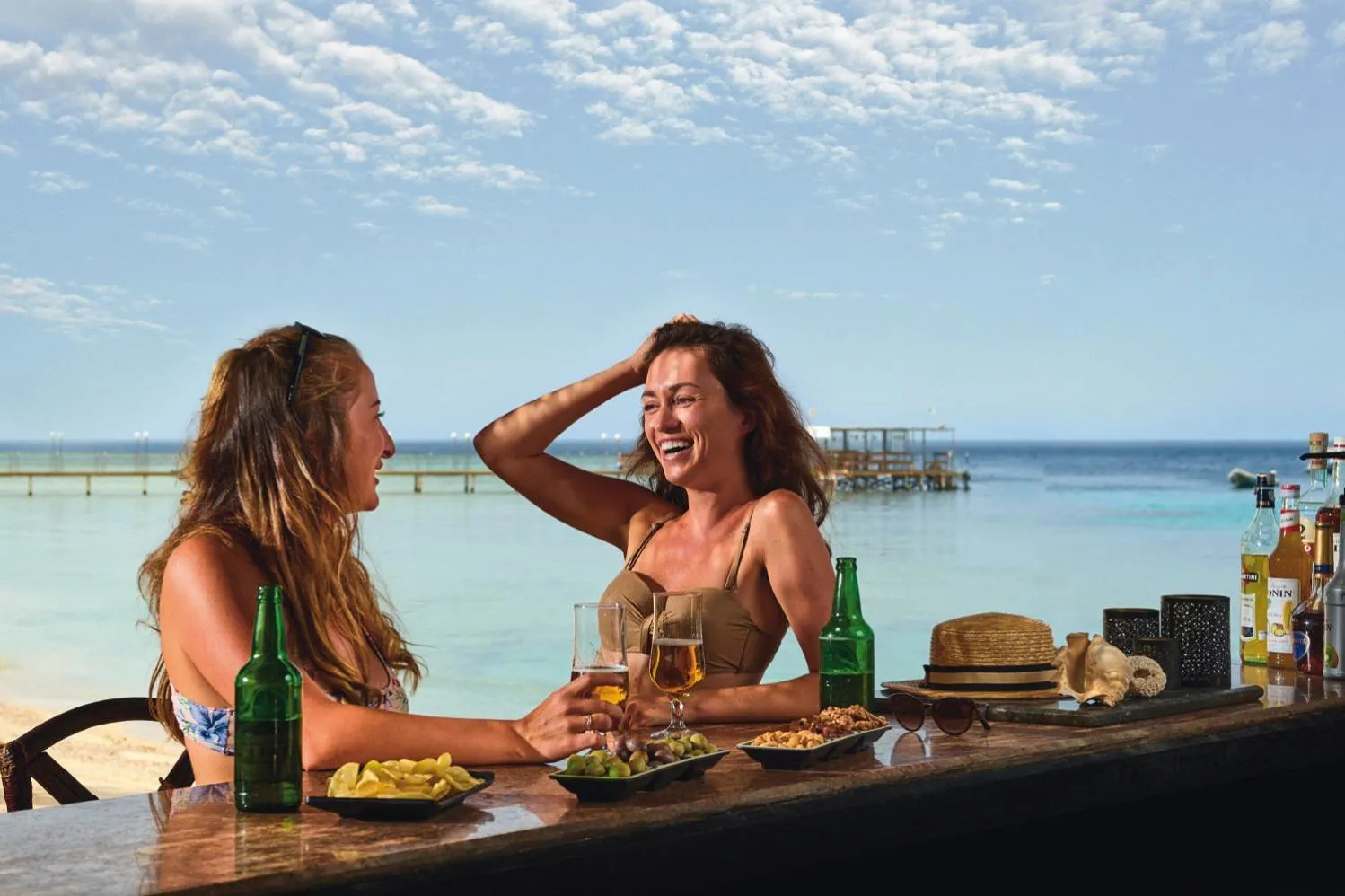 Deux femmes sourient en sirotant des boissons au comptoir d'un bar en bord de mer.