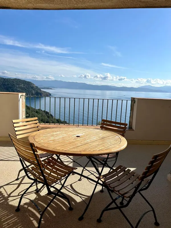 Une table ronde en bois et des chaises en métal sur une terrasse ensoleillée avec vue sur la mer et les montagnes.