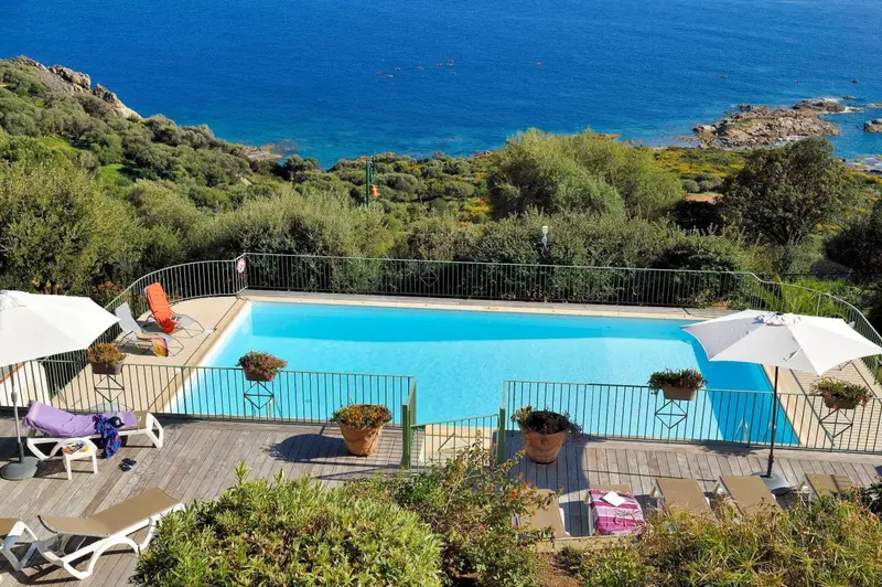 Une piscine rectangulaire avec des transats et des parasols surplombe une crique rocheuse.