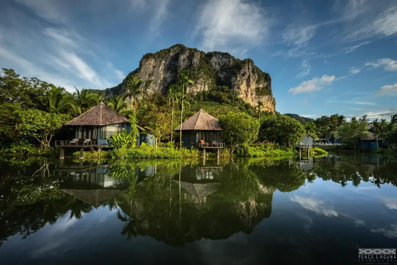 Des bungalows sur pilotis se reflètent dans l'eau calme devant une falaise verdoyante.