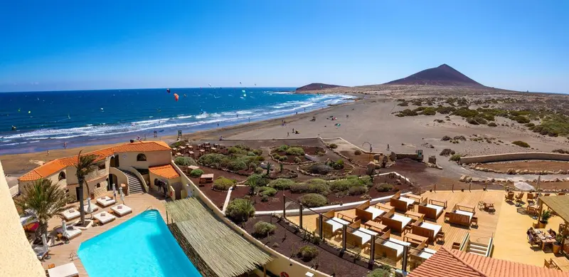 Vue aérienne d'un complexe hôtelier avec piscine et transats, face à une plage et une mer agitée.
