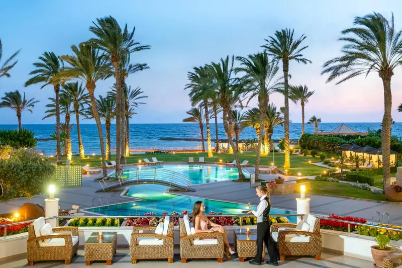 Une piscine bordée de chaises longues et de végétation tropicale, avec un serveur apportant des boissons à des clients.