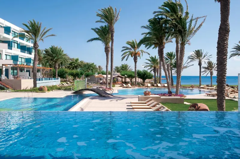 Vue d'une piscine bleue bordée de transats, avec un bâtiment et des palmiers en arrière-plan.