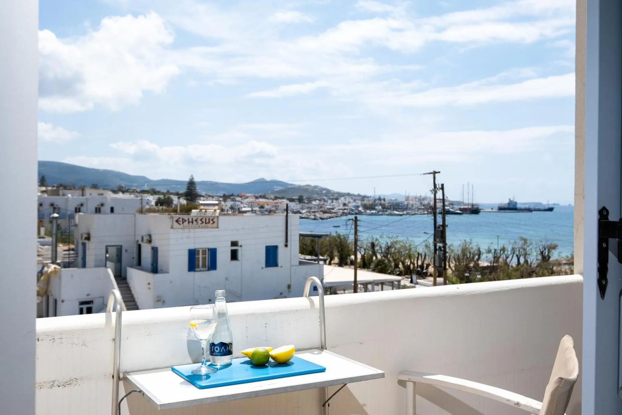 Une table avec des boissons et des fruits est posée sur un balcon avec vue sur la mer et la ville.
