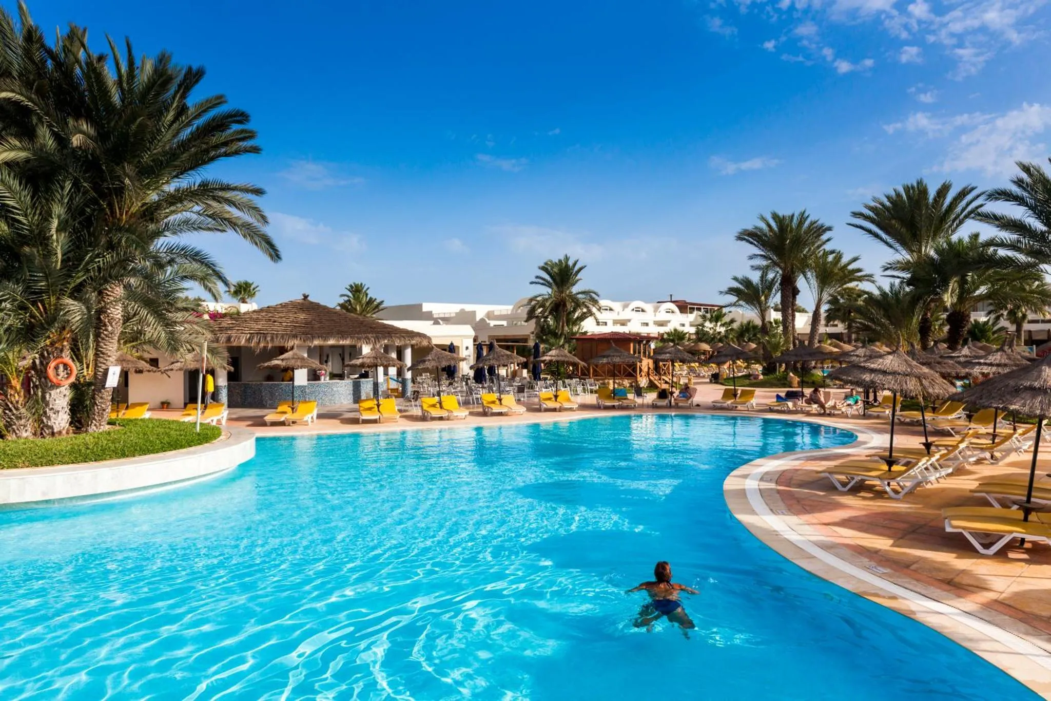 Une piscine turquoise bordée de transats sous des palmiers, avec une personne nageant au centre.
