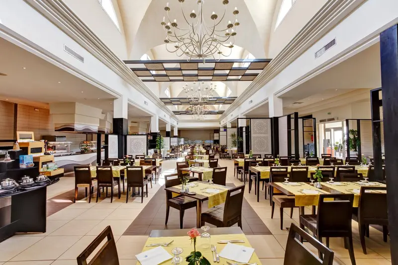 Une grande salle lumineuse est aménagée en restaurant avec des tables dressées et un buffet.