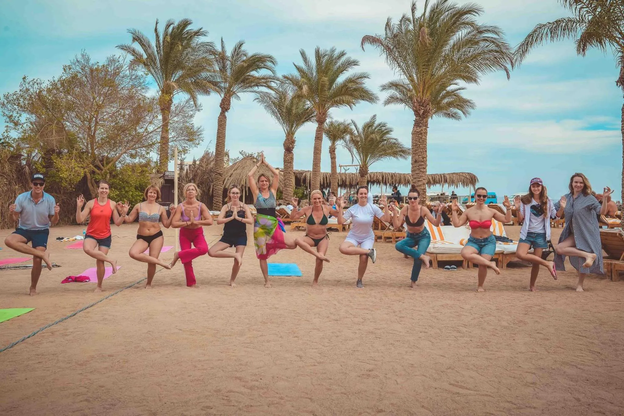 Groupe de personnes pratiquant le yoga sur une plage de sable fin, avec des palmiers en arrière-plan.