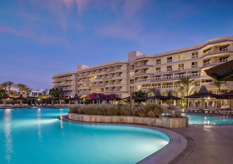 Bâtiment blanc illuminé au crépuscule surplombant une piscine turquoise et des palmiers.