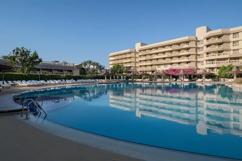 Une grande piscine rectangulaire reflète un bâtiment beige à plusieurs étages sous un ciel bleu clair.