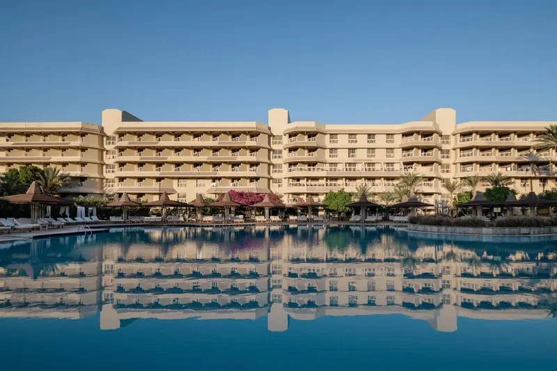 Bâtiment beige à plusieurs étages surplombant une piscine turquoise et des transats.