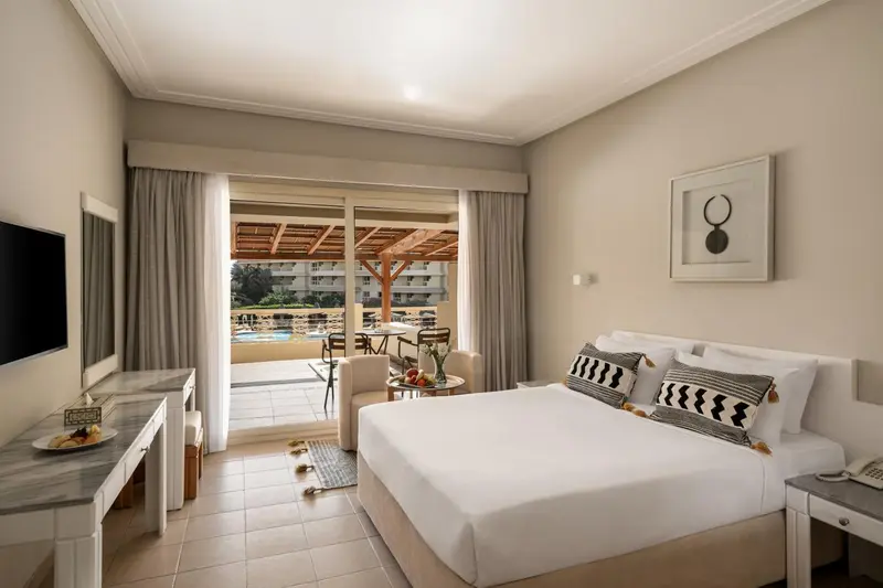 Chambre lumineuse avec un grand lit et une terrasse donnant sur une piscine.