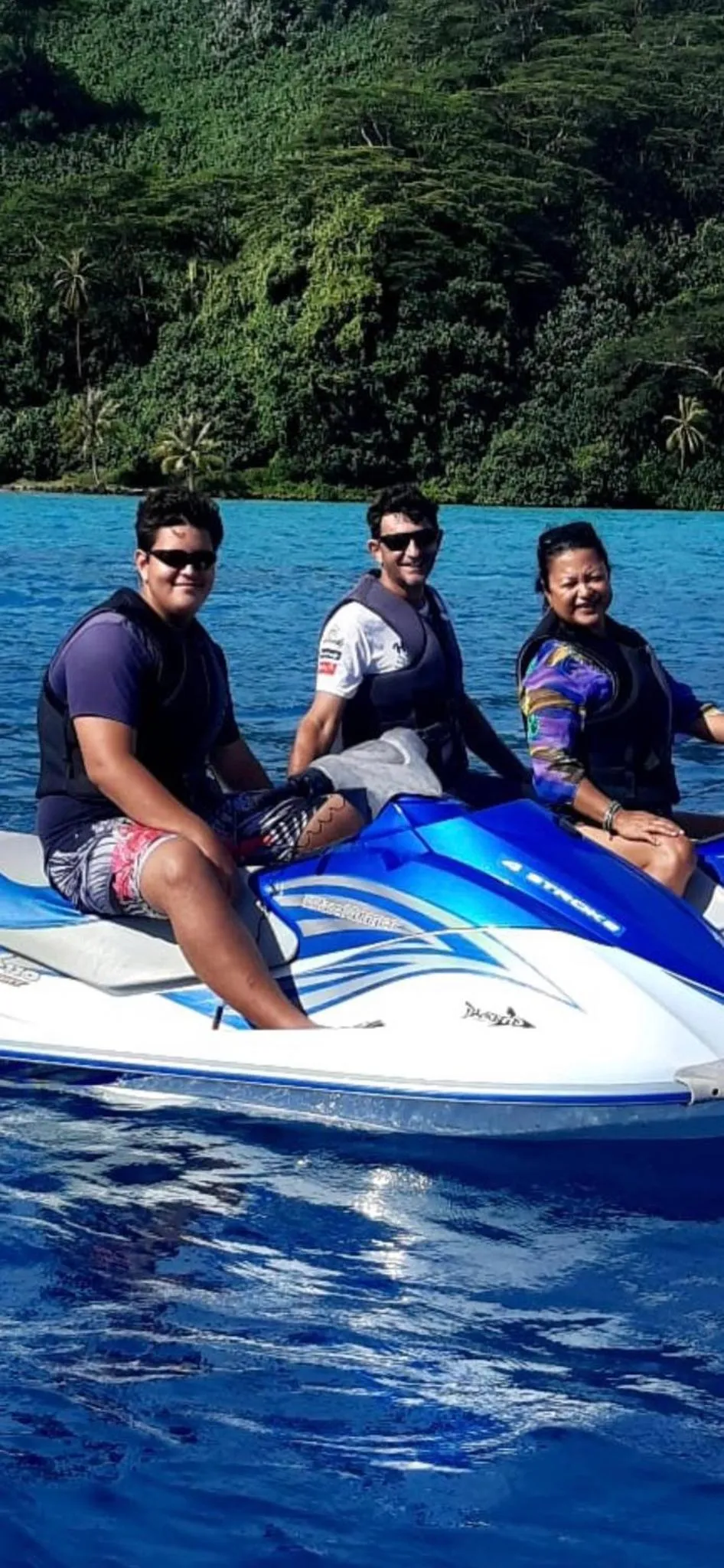 Trois personnes sourient sur un jet ski bleu au bord de l'eau turquoise.