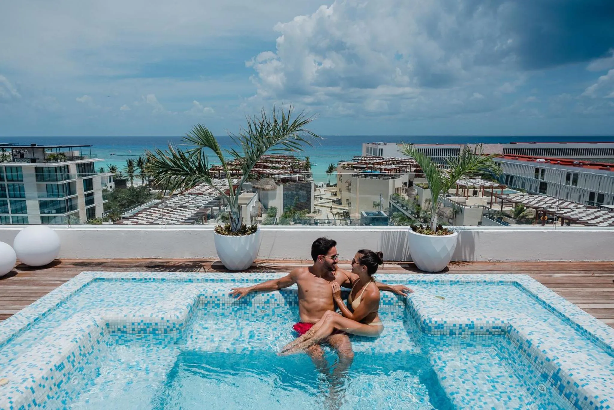 Un couple se détend dans un jacuzzi sur une terrasse avec vue sur la mer et les bâtiments environnants.