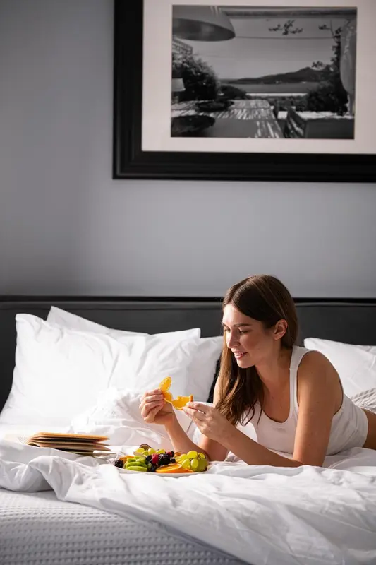 Une femme souriante déguste des fruits frais allongée sur un lit blanc.