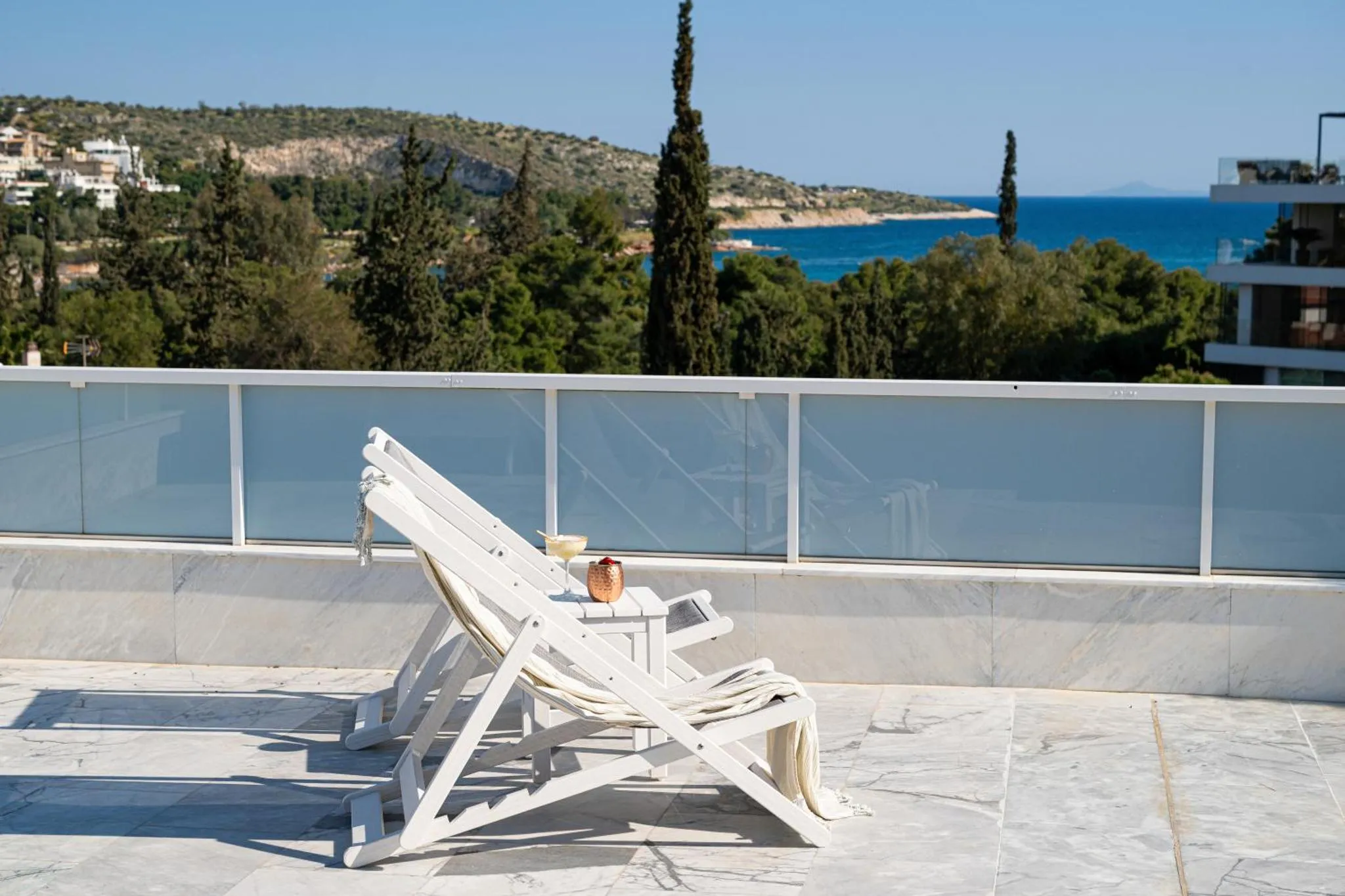 Transats blancs sur une terrasse carrelée avec vue sur la mer et la végétation.