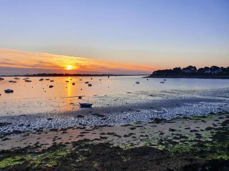 Coucher de soleil sur une étendue d'eau avec des bateaux amarrés et une plage au premier plan.