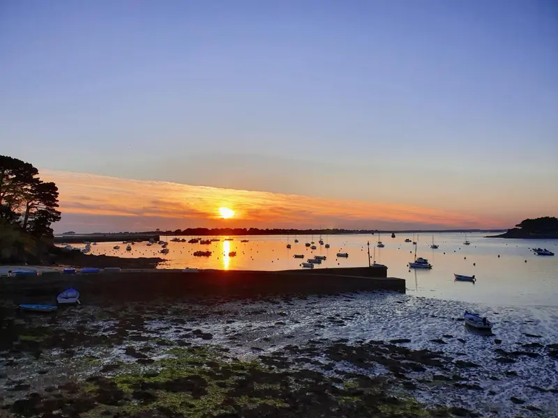 Coucher de soleil sur une baie avec des bateaux amarrés et une plage au premier plan.