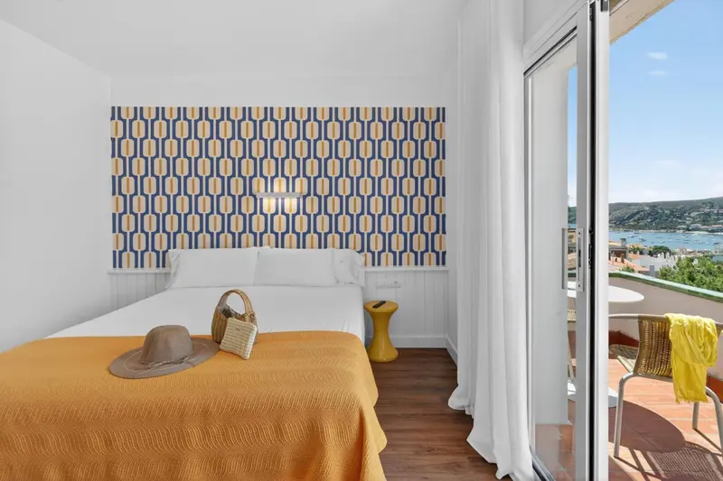 Une chambre lumineuse avec un lit recouvert d'une couverture orange et un chapeau de paille posé dessus.