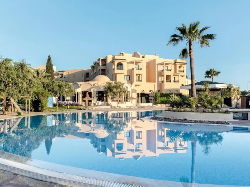Bâtiment beige à plusieurs étages avec piscine au premier plan et palmiers.