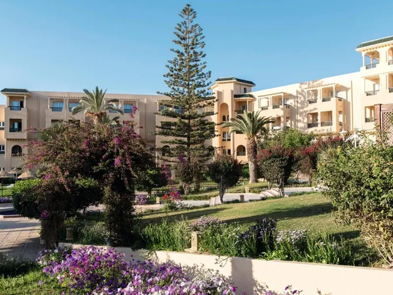 Bâtiment beige à plusieurs étages entouré d'une végétation luxuriante et d'un jardin fleuri.