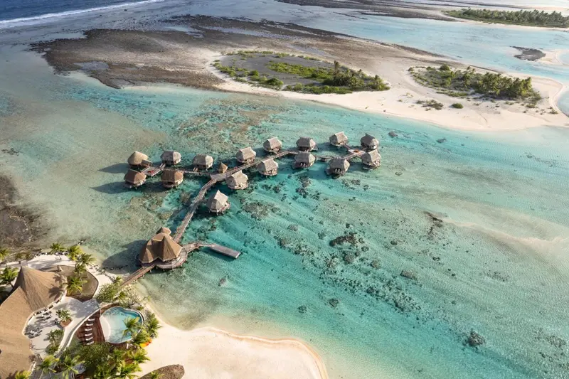 Vue aérienne d'un complexe hôtelier avec des bungalows sur pilotis au-dessus d'un lagon turquoise.