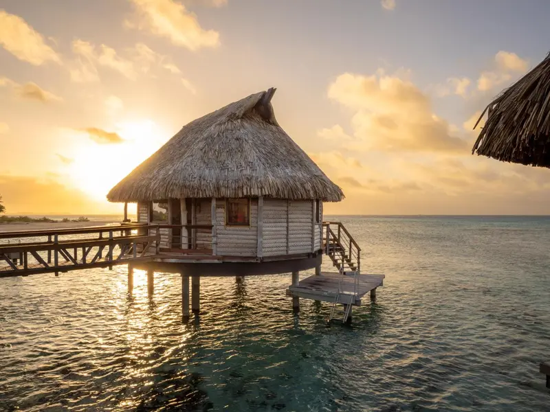 Une cabane sur pilotis au-dessus d'une eau turquoise au coucher du soleil.