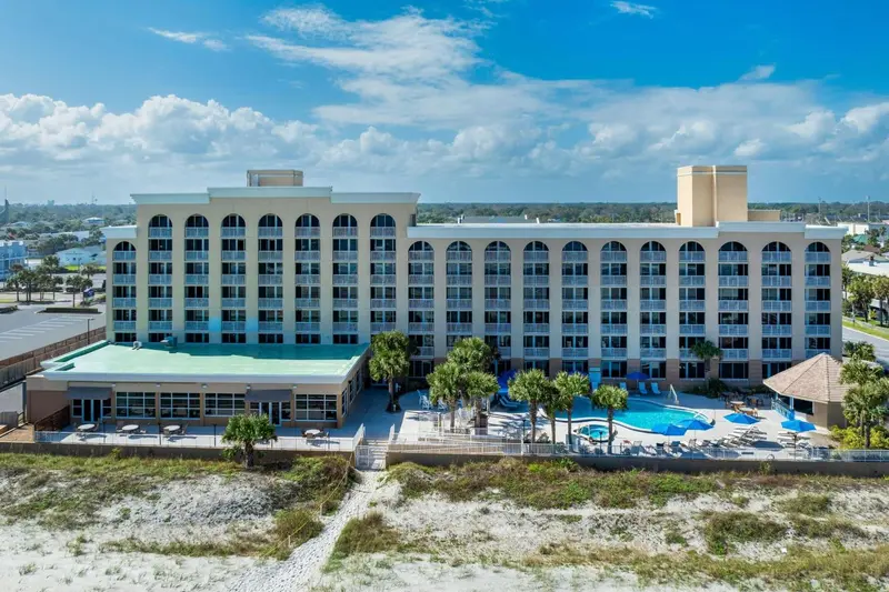 Bâtiment beige à plusieurs étages avec des balcons donnant sur une plage de sable blanc et une piscine.