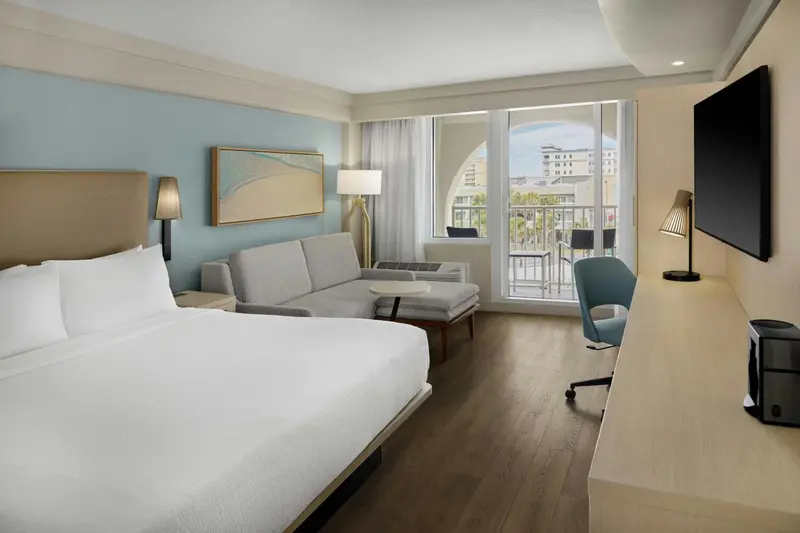 Chambre lumineuse avec un lit blanc et un canapé gris face à une baie vitrée.